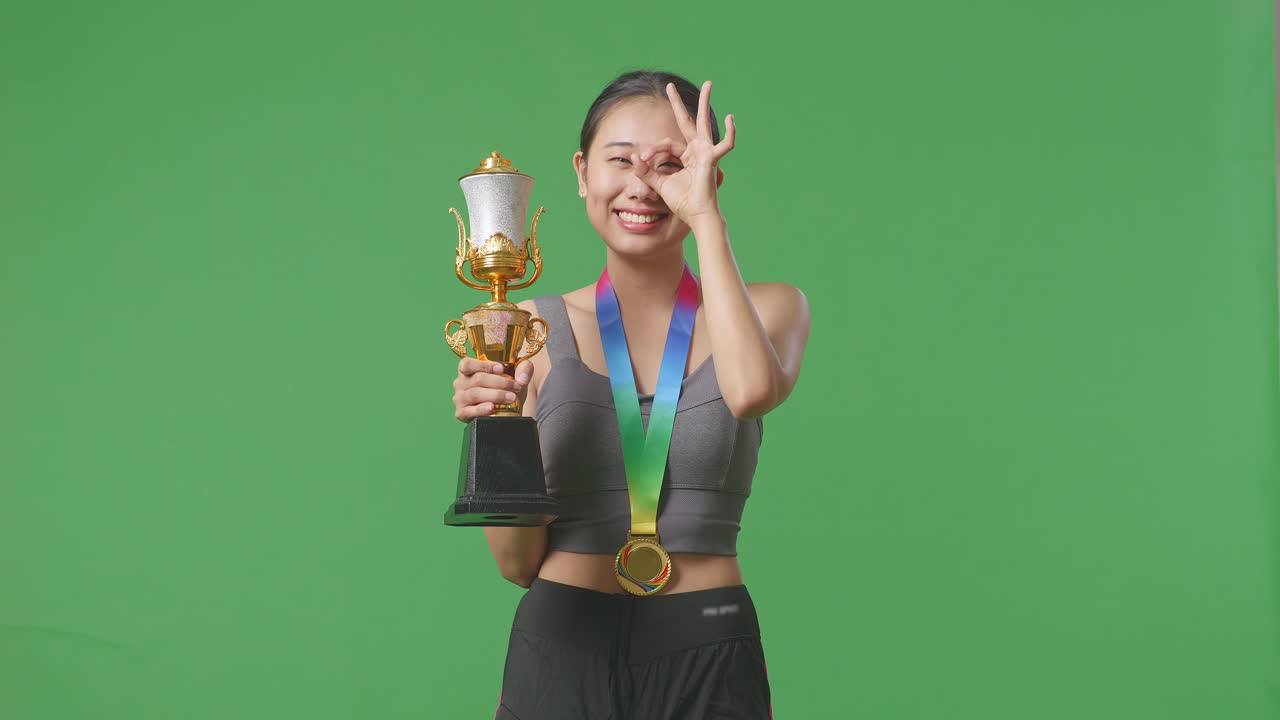 금메달과 트로피를 들고 있는 아시아 여성은 오케이 손으로 눈을 감고 카메라에 미소를 지으며 스튜디오의 녹색 스크린 배경에서 첫 번째 우승자로 나타납니다.