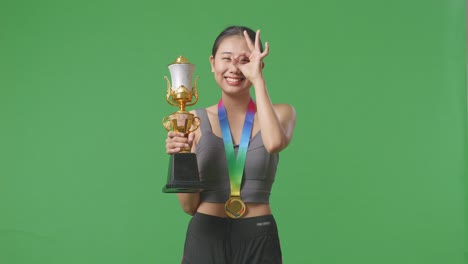 asian woman with a gold medal and trophy showing okay hand sign over eye and smiling to camera as the first winner on green screen background in the studio