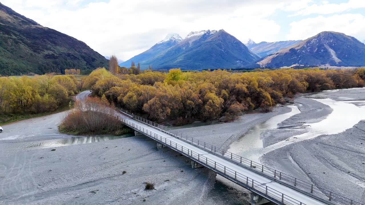 Aerial view of a car crossing a bridge over a riverbed with mountains in the background, under bright daylight