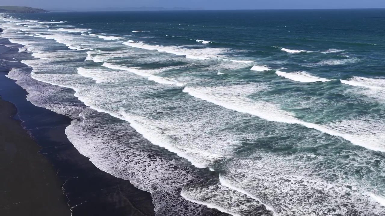 Vista aerea di onde oceaniche che si infrangono su una lunga spiaggia di sabbia nera, in piena luce del giorno