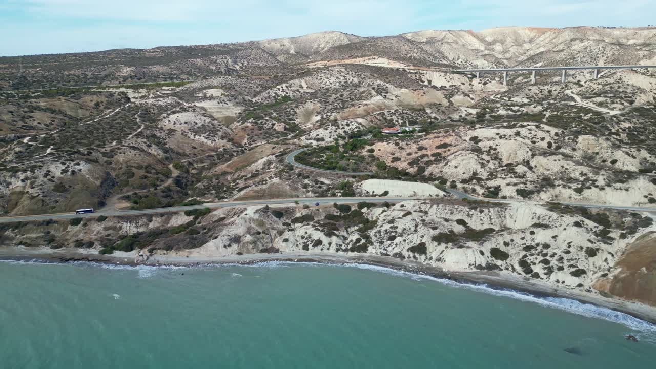 Coastal aerial of Petra tou Romiou with rocky shoreline, hills and turquoise clear sea, panoramic pullback from road