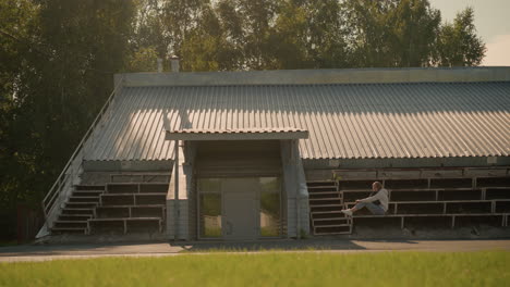 young woman seated alone on rustic stadium bleachers under metal roof, surrounded by greenery and soft shadows, in a contemplative mood