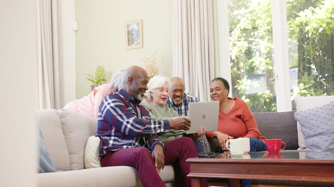 Happy diverse senior friends using laptop together in sunny living room, copy space, slow motion