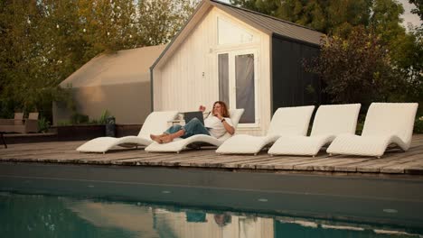 a curly-haired guy in glasses is lying on a white deckchair near the sunbeds by the pool and working on a laptop. rest in the country house