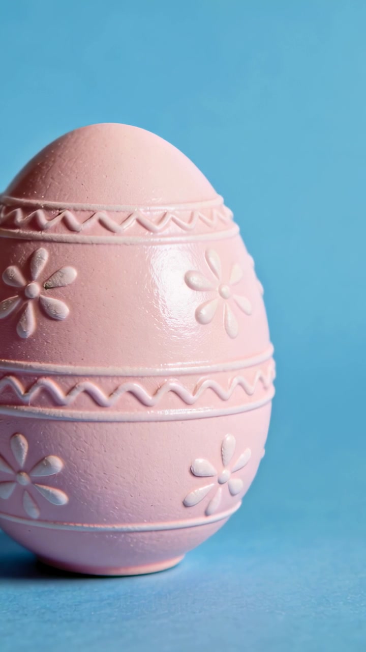 Shifting frame nudging camera tightening pink egg on blue set, centering daisy motifs, zigzag bands