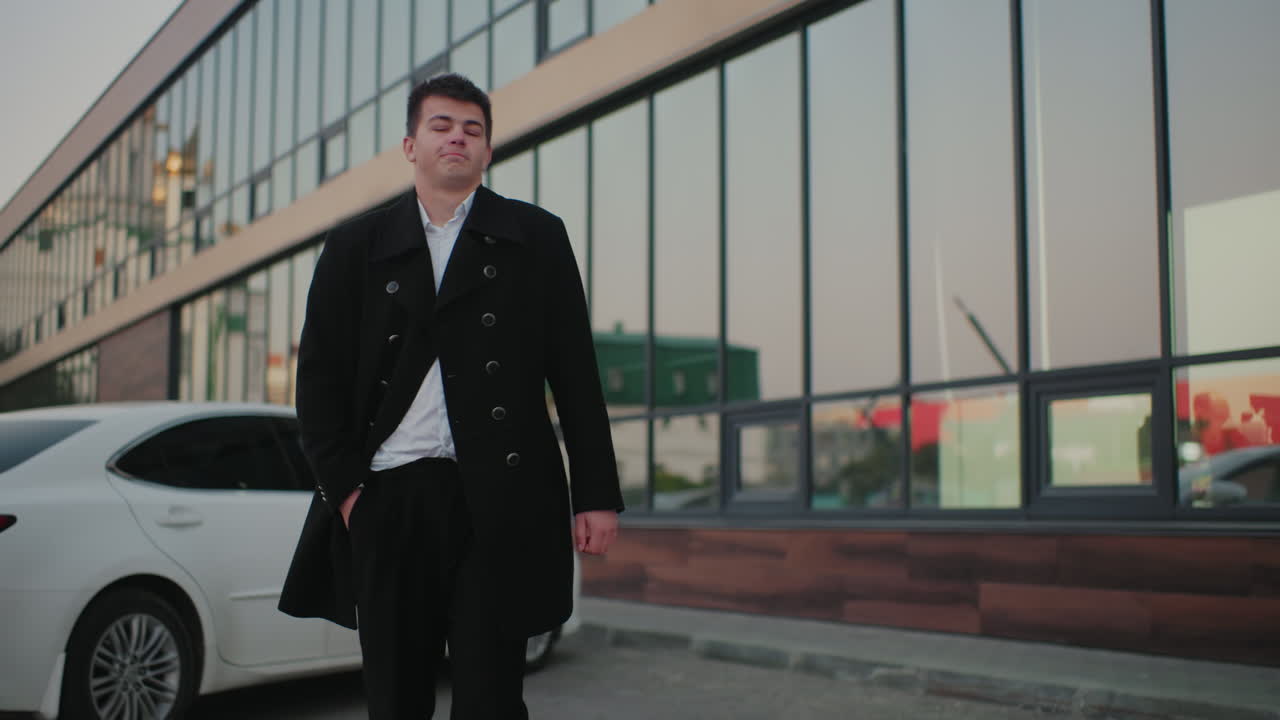 검은 코트를 입은 남자가 코트를 조정하고, 주머니에 손을 넣고, 도시 비즈니스 환경을 통해 웅장하게 고, 사무실 건물과 주차된 차와 함께 전문적인 기