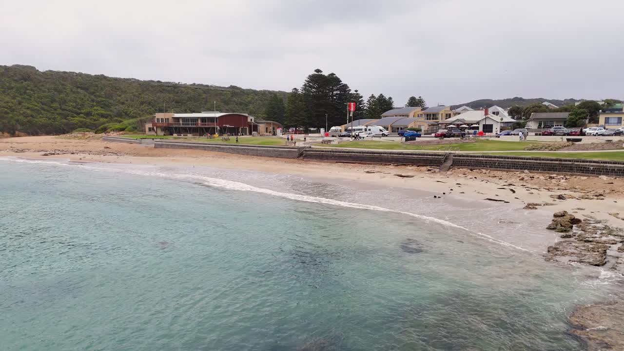 Drone footage captures Port Campbell's coastline, showcasing clear waters, sandy beaches, and nearby buildings under overcast skies