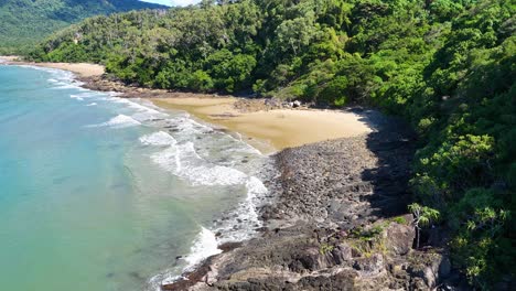 drone footage rises over a deserted tropical beach and lush rainforest, capturing clear turquoise water, rocky shoreline, and bright natural daylight in port douglas, australia