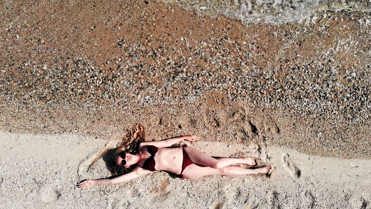 Young woman sitting on the beach sand near water and relaxing. Top view. Slow motion