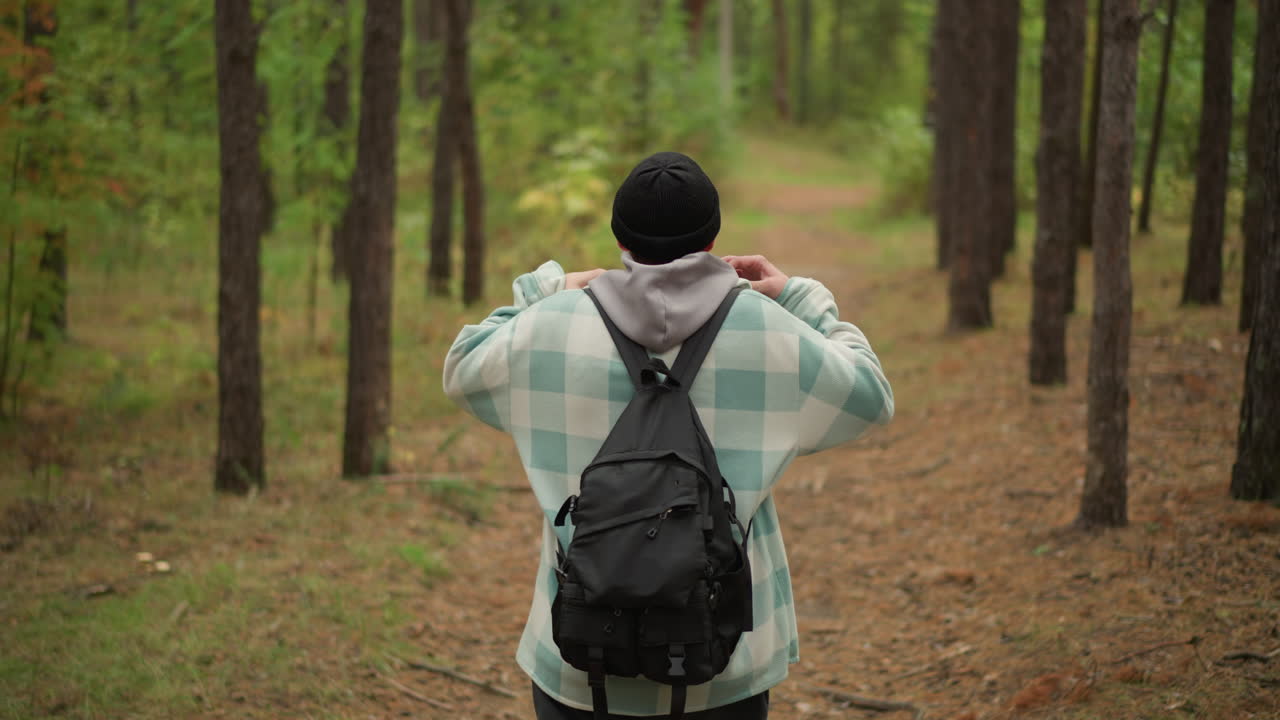 Back view white hiker walking forest trail among tall pines, backpack and beanie, hoodie, slow steady pace, soft autumn light, path fading into distance, contemplative mood, nature escape