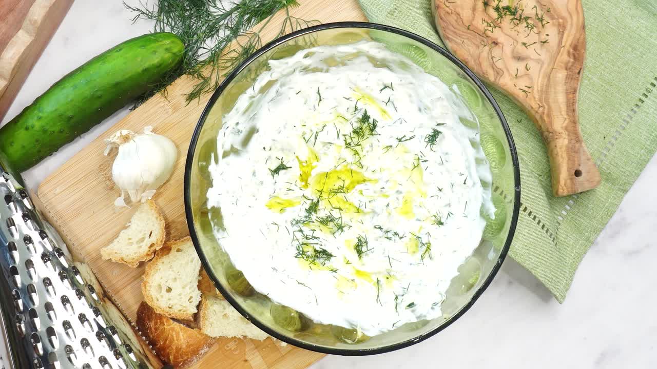 Making Tzatziki Greek or Mediterranean sauce or dip for a party. Dip or sauce in a green bowl on a wooden cutting board with dill, green napkin and a grater. Greek Tzatziki sauce for all foods.