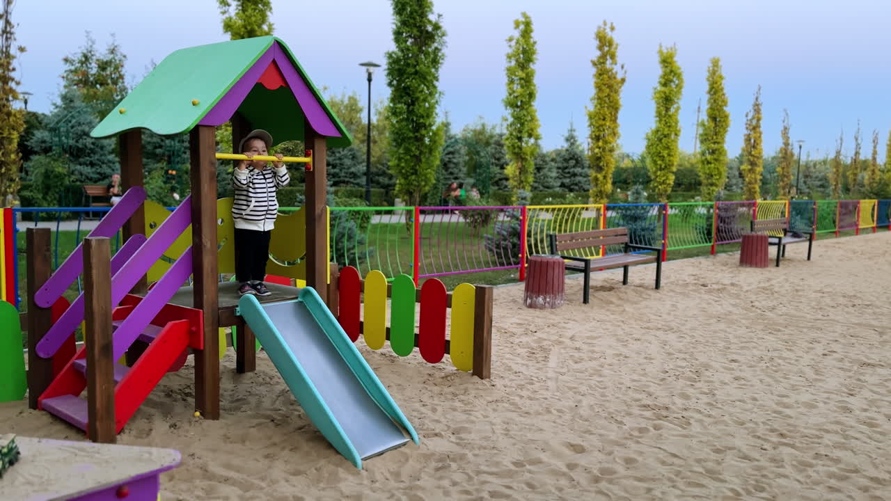 Lovely toddler in stripy jacket hanging on the horizontal bar at playground. Happy kid jumps cheerfully on top of the slide.