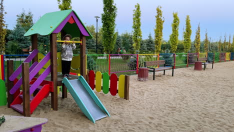 lovely toddler in stripy jacket hanging on the horizontal bar at playground. happy kid jumps cheerfully on top of the slide.