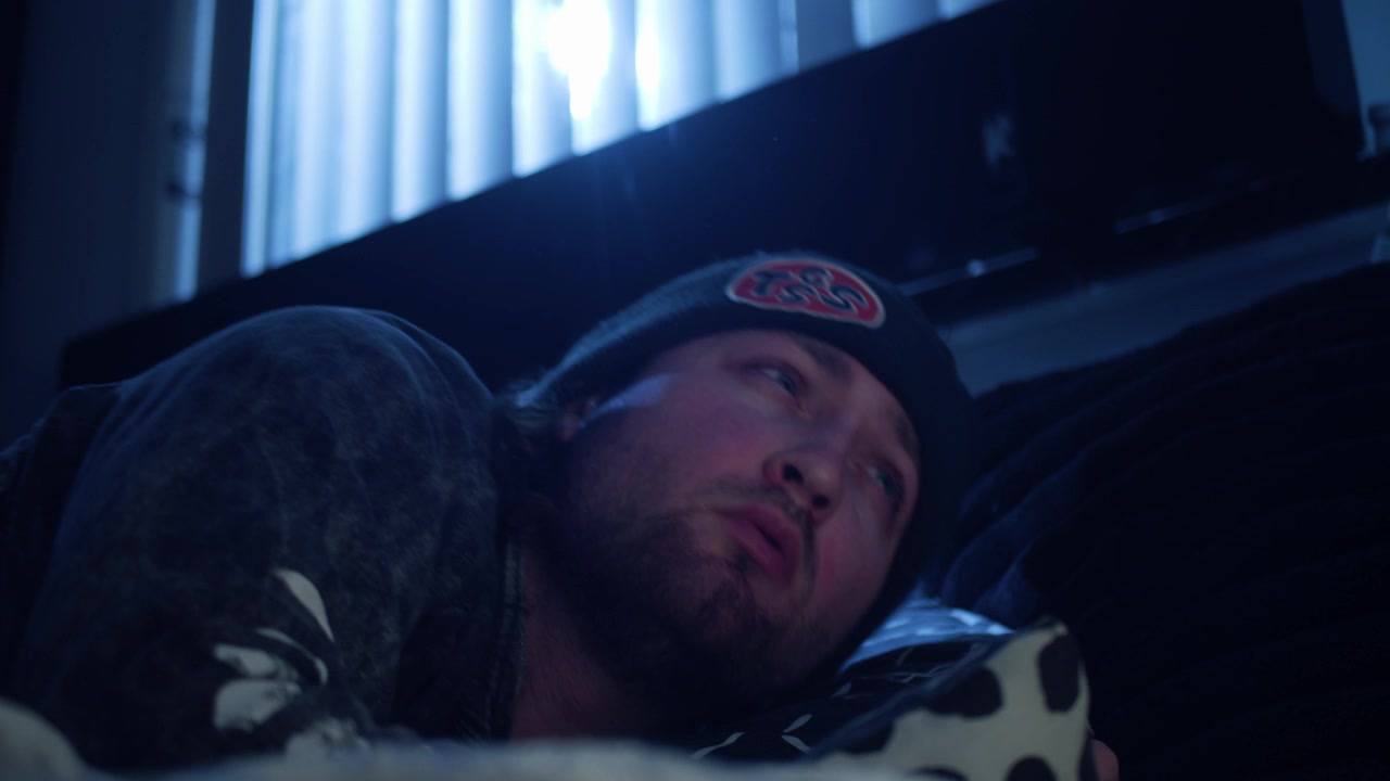 Ripresa cinematografica in 4K di un uomo che dorme in una camera da letto illuminata dalla luna blu, prima di girarsi, reagire a una luce intensa e sedersi con gli occhi socchiusi.