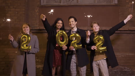 group of friends wearing elegant clothes holding sparklers and 2022 numbers golden ballons in the street after the new year's party