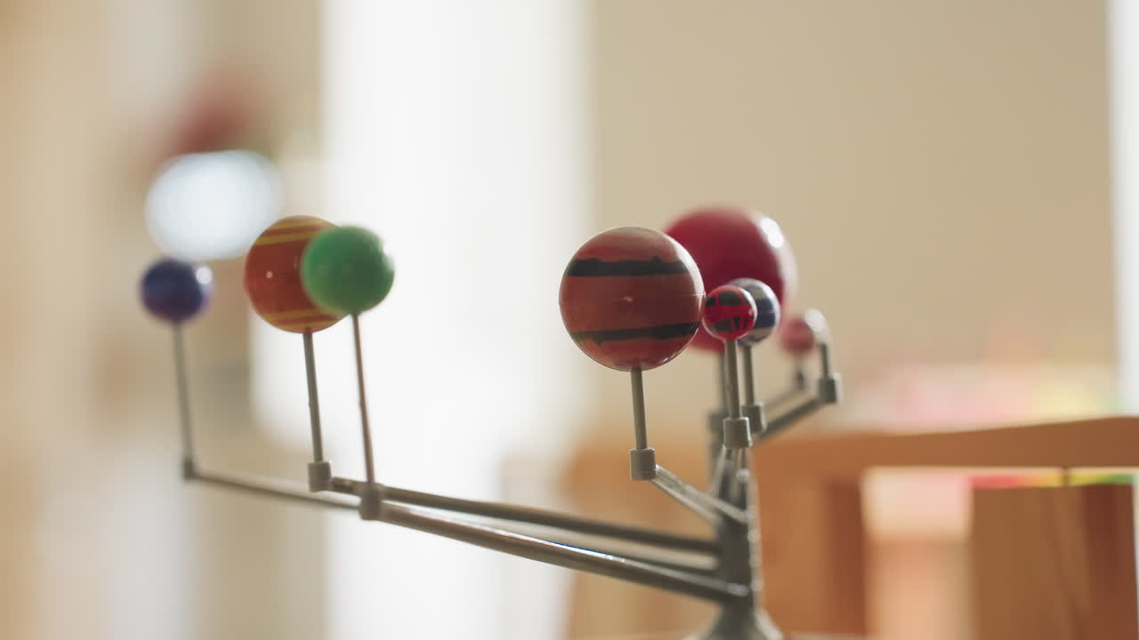 Close up of colorful solar system model showing nine planets mounted on metal rods, standing on wooden surface in bright educational room with blurred warm background