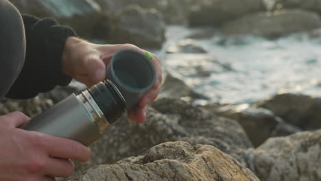 man screws a cup on thermos as sea waves wash over rocks in background during golden hour