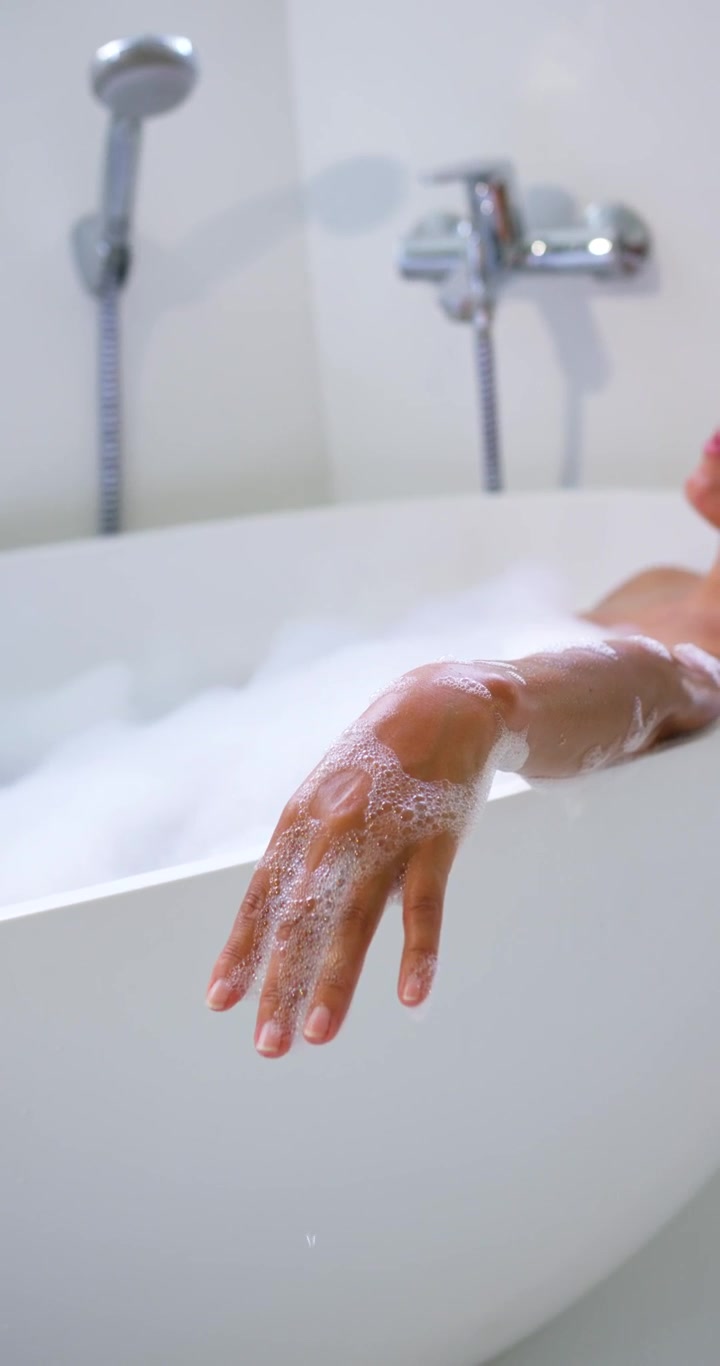 Vertical video: Leaning woman keeping arm over tub rim at home, relaxing with foam, tap, copy space