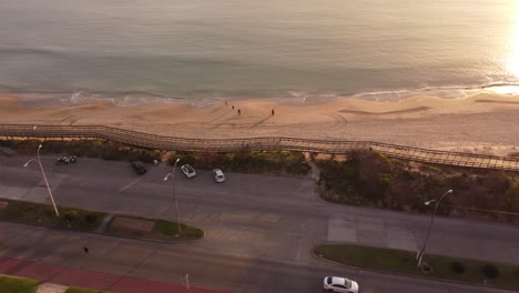 Vista-Aérea-De-La-Gente-Caminando-Por-La-Playa-De-Arena-Y-La-Costa-De-Punta-Del-Este-Y-Disfrutando-De-La-Puesta-De-Sol-Dorada---Camiones-Conduciendo-Por-La-Carretera-Costera-Por-La-Noche