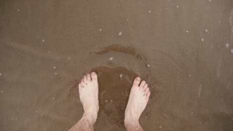 a mans feet stood on the beach as the tide comes in and water laps at the shore