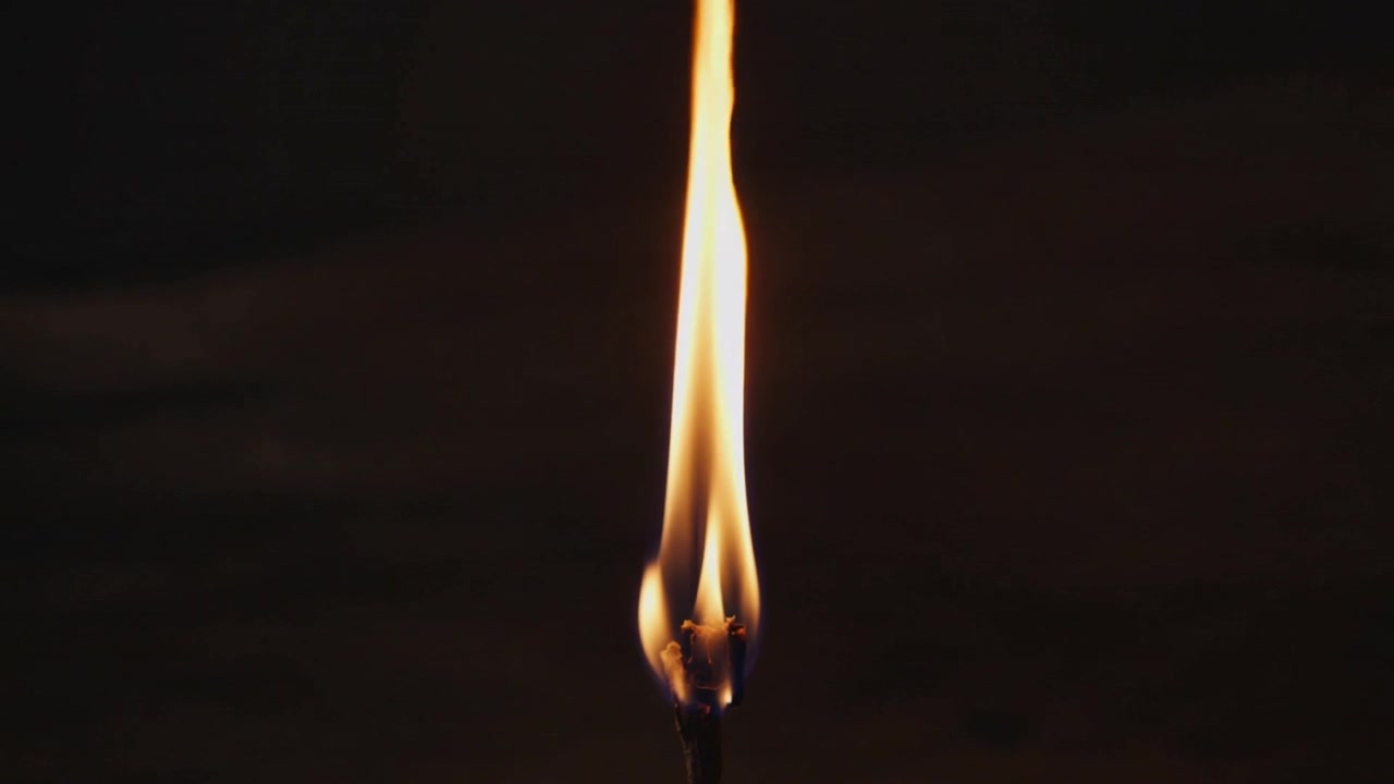 Flickering flame atop charred stick stretching upward while burning in dim studio, dark wood