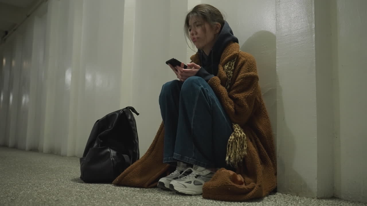 A girl in a brown coat, jeans, and white shoes sits bent over in an underpass tunnel, holding her phone with an unhappy expression.The scene conveys a strong sense of loneliness and emotional struggle