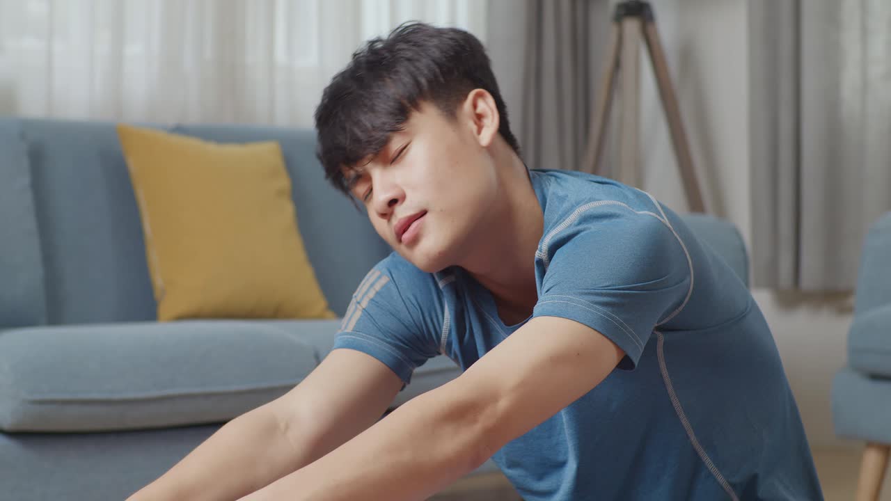 운동복을 입은 젊은 아시아 운동선수 남성이 요가 매트에 앉아서 집에서 햄스트링 스트레칭을 하고 있다.
