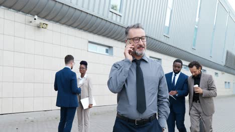 hombre de negocios canoso con gafas y hablando por teléfono mientras camina por la calle