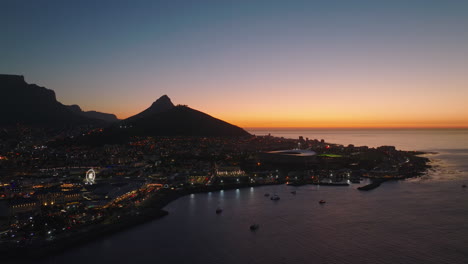 Forwards-fly-above-water-surface.-Waterfront-and-city-at-dusk,-silhouette-of-mountain-peak-against-colourful-sky.-Cape-Town,-South-Africa