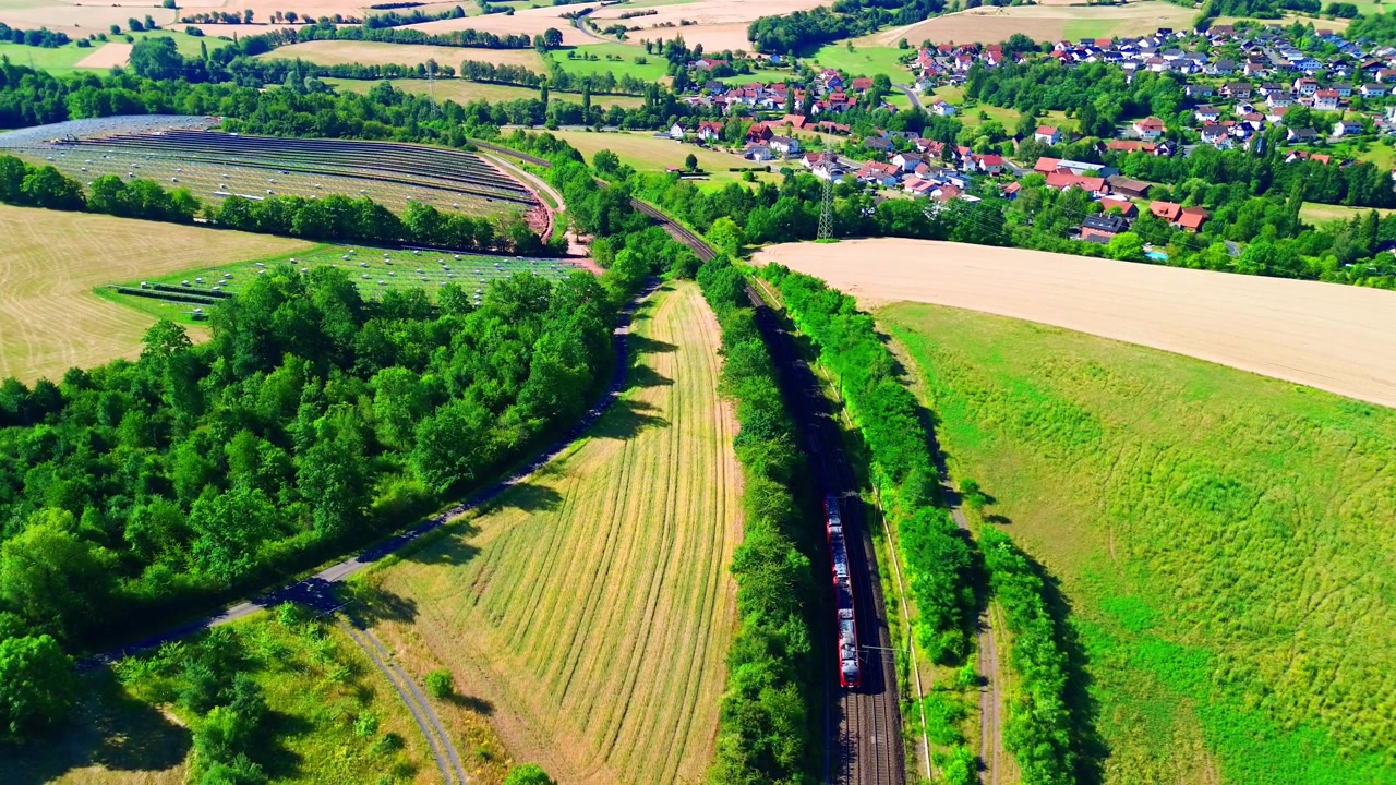 Luchtfoto van een rode regionale passagierstrein op een geëlektrificeerde lijn tussen een zonnepark op een heuvel en een Duits dorp omringd door zomerse velden en bosland