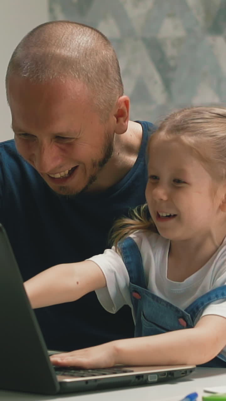 closeup slow motion dad with little daughter laugh, sitting at table, look at computer screen, smiling, communicating by video link, pointing finger enthusiastically, very interested home education