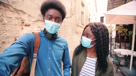 african american woman and man wearing masks and waving at camera while smiling in the street