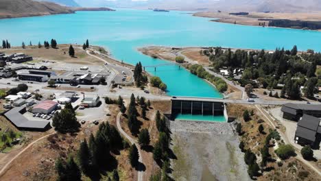 Aerial-View-of-Lake-Tekapo-Town,-Traffic-on-Bridge-Above-Control-Gates-to-River-Outflow-and-Aqua-Blue-Water,-Drone-Shot