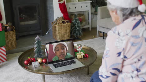 mujer caucásica senior agitando y usando computadora portátil para una videollamada de navidad con un niño en la pantalla