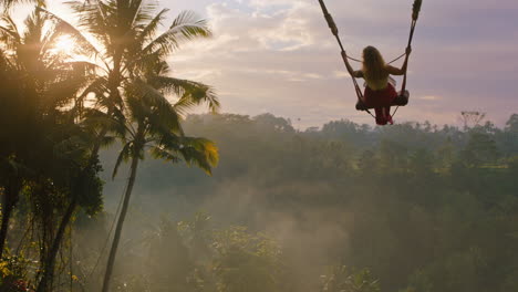 travel woman swinging over jungle at sunrise enjoying exotic vacation sitting on swing with sun flare shining through palm trees in tropical rainforest holiday lifestyle freedom