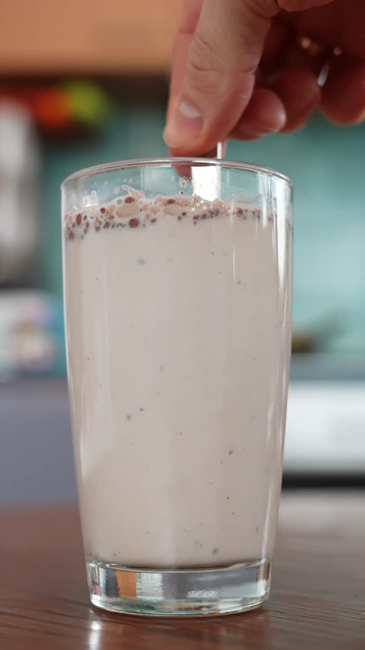 Una mano mescola latte e cioccolato fatto in casa in un bicchiere alto su un tavolo di legno, cremosa bevanda al cacao con schiuma e granelli, sfondo tenue della cucina, primo piano verticale