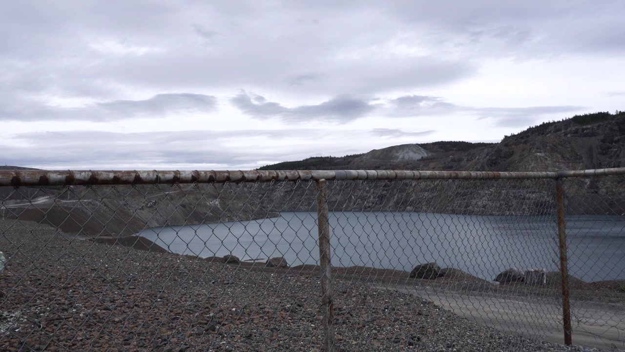 Un'ampia veduta di una miniera a cielo aperto allagata, ripresa da dietro una recinzione di sicurezza a rete metallica su una sponda rocciosa, con pareti di scavo aspre e un cielo coperto