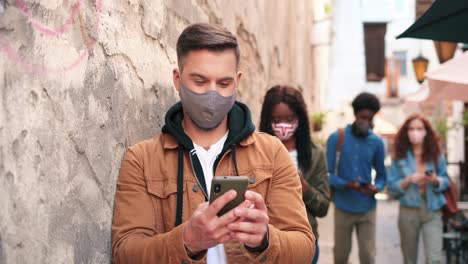 vista de cerca del hombre caucásico con máscara y usando un teléfono inteligente apoyado en una pared en la calle