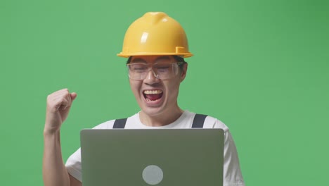 close up of asian man worker wearing goggles and safety helmet looking at the laptop then screaming goal celebrating while standing in the green screen background studio