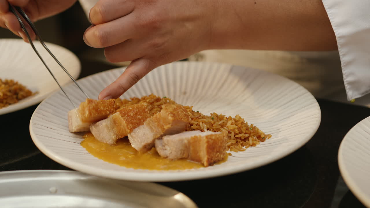 A chef uses tongs to place a piece of cooked pork on a bed of sauce with rice on the side