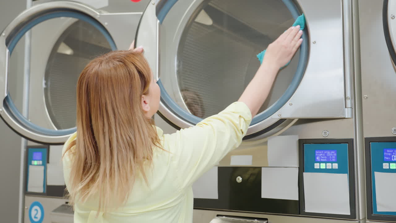 Laundress approaches industrial washer in modern laundry facility, opening stainless steel door to begin cleaning transparent surface, preparing machine for use with attention to hygiene