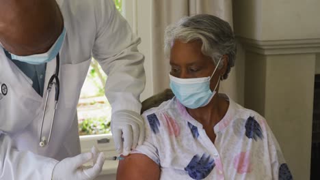 african american senior male doctor injecting covid-19 vaccine into african american woman at home