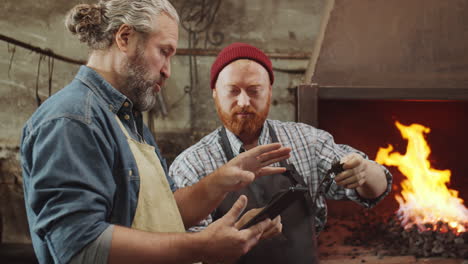 two blacksmiths discussing metal details and using tablet in smithy