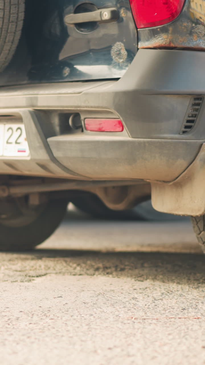 Primer plano de la rueda sucia de un todoterreno en el pavimento, llanta con salpicaduras de barro y banda de rodadura desgastada cerca del parachoques trasero y matrícula visible, conductor inspeccionando el neumático en busca de un pinchazo, mecánico de carretera acercándose con un juego de herramientas