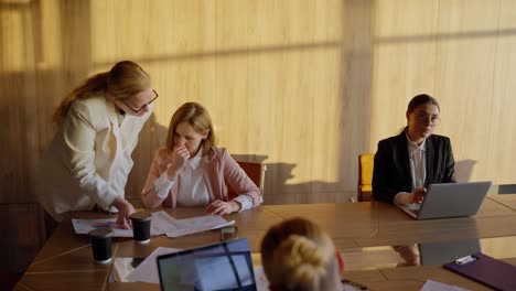 side view of a group of businesswoman in business suits sitting at a wooden table in a sunny office communicating and working on laptops
