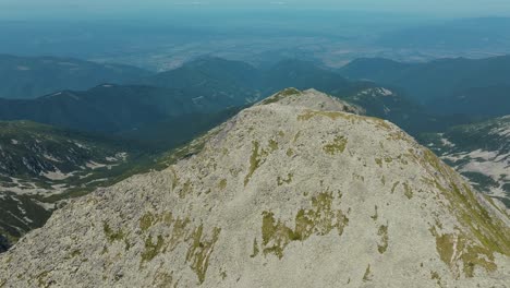 Rocky mountain tall peak surrounded by mountain ranges, on a clear sunny day with blue skies and vibrant green grass, drone tracking shot