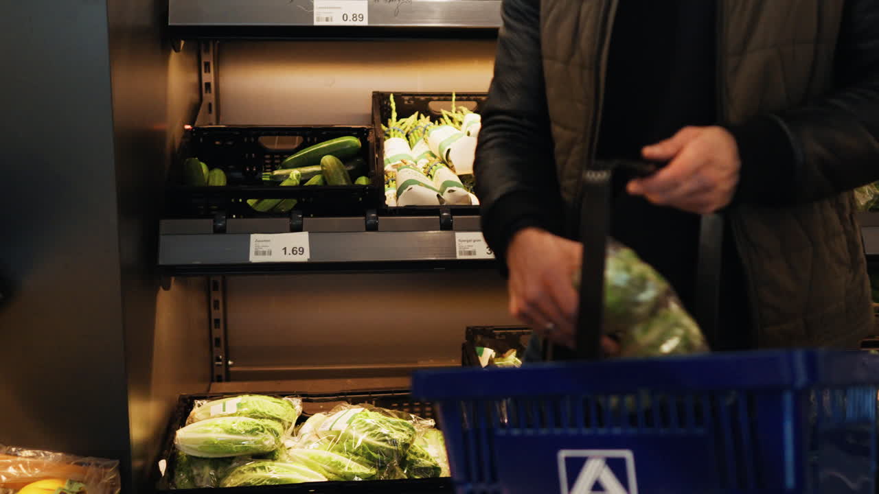 A man selects fresh green salad leaves from the produce section and places them into his basket while shopping in a grocery store.