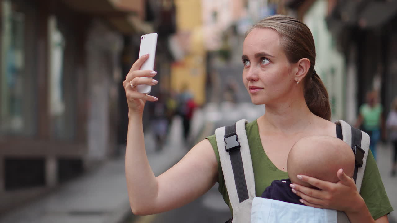 With her baby in a kangaroo backpack, a young woman photographs her adventures on a mobile phone during the journey. She walks and frequently glances at the phone's screen