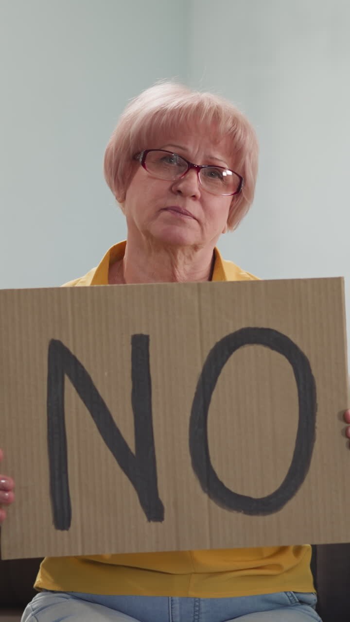 Upset senior woman holds placard with word NO sitting on sofa in living room slow motion. Protest against social and political events. Negative decision