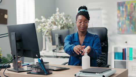 an attractive woman in a shirt and glasses comes to work, sits down at desk, disinfects her hands with a liquid before clicking fingers on the computer keyboard, company health and safety
